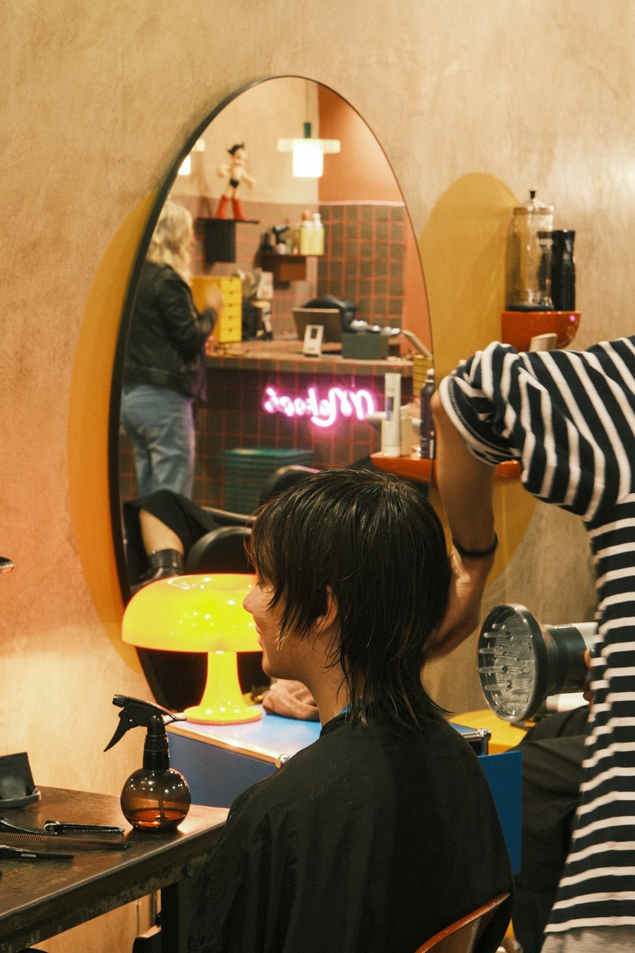 A stylist blow-drying a client's freshly cut hair in front of a round salon mirror